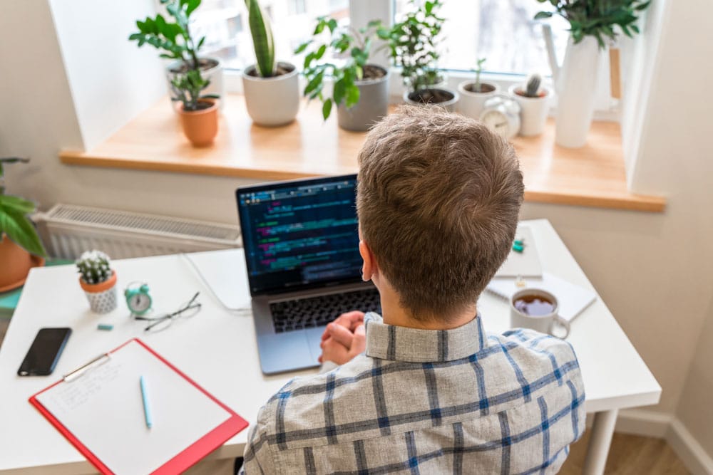 Чоловік сидить за столом біля вікна та програмує на ноутбуці у домашньому офісі.